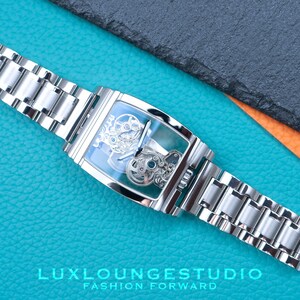 Puede incluir: Un reloj plateado con una esfera cuadrada y un movimiento esqueletizado. El reloj tiene una correa de metal plateado y est&aacute; sobre una superficie turquesa.