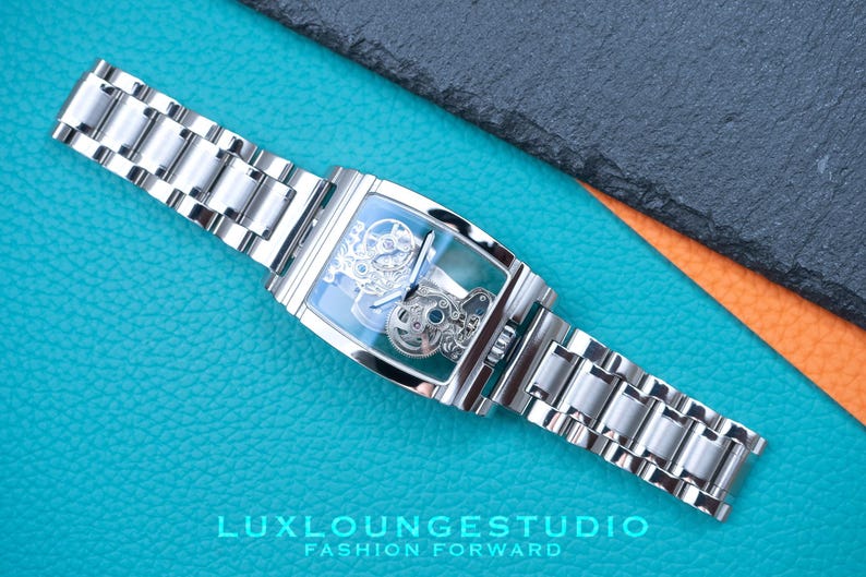 Puede incluir: Un reloj plateado con una esfera cuadrada y un movimiento esqueletizado. El reloj tiene una correa de metal plateado y est&aacute; sobre una superficie turquesa.