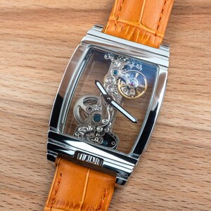 May include: A rectangular, silver-toned wristwatch with a transparent face, showcasing the intricate internal mechanisms. The watch has a light orange leather band. The watch is set against a wooden background.