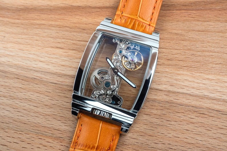 May include: A rectangular, silver-toned wristwatch with a transparent face, showcasing the intricate internal mechanisms. The watch has a light orange leather band. The watch is set against a wooden background.