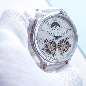 Pu&ograve; includere: Un orologio argentato con quadrante bianco e indici delle ore in numeri romani. L'orologio ha una complicazione di fase lunare e un doppio tourbillon. Il quadrante dell'orologio presenta anche il testo "AILANG" e "DOUBLE TOURBILLON AUTOMATIC".