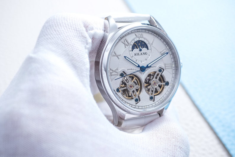 Pu&ograve; includere: Un orologio argentato con quadrante bianco e indici delle ore in numeri romani. L'orologio ha una complicazione di fase lunare e un doppio tourbillon. Il quadrante dell'orologio presenta anche il testo "AILANG" e "DOUBLE TOURBILLON AUTOMATIC".