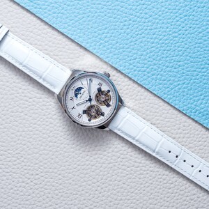 Pu&ograve; includere: Un orologio da polso bianco con quadrante argentato e cinturino in pelle bianca. L'orologio ha un indicatore delle fasi lunari, una finestrella per la data e un movimento scheletrato.