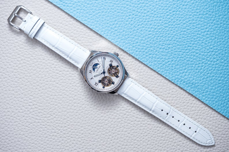 Pu&ograve; includere: Un orologio da polso bianco con quadrante argentato e cinturino in pelle bianca. L'orologio ha un indicatore delle fasi lunari, una finestrella per la data e un movimento scheletrato.