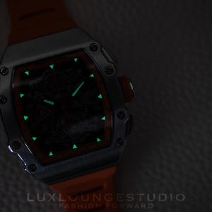 Peut inclure: Une montre de luxe avec un bo&icirc;tier argent&eacute; et un bracelet orange. Le cadran de la montre est sombre avec des marqueurs et des aiguilles verts brillants. L'image comprend le texte "LUXLOUNGESTUDIO FASHION FORWARD".