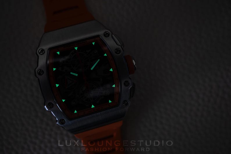 Peut inclure: Une montre de luxe avec un bo&icirc;tier argent&eacute; et un bracelet orange. Le cadran de la montre est sombre avec des marqueurs et des aiguilles verts brillants. L'image comprend le texte "LUXLOUNGESTUDIO FASHION FORWARD".