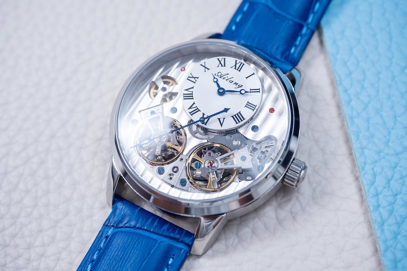 May include: A silver-toned wristwatch with a blue leather band. The watch face is white with Roman numeral hour markers and a skeletonized movement visible through the clear case back. The watch face also features the word "Aidlang" in black.