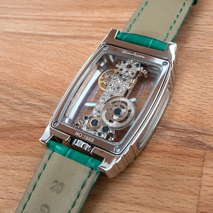 May include: A silver rectangular wristwatch with a transparent face, revealing intricate internal gears and jewels. The watch features a green leather strap. The text "Ginstoria" is visible on the watch face, and the number "NO. 1688" is also visible.