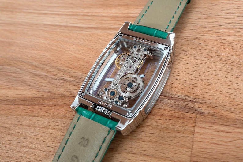 May include: A silver rectangular wristwatch with a transparent face, revealing intricate internal gears and jewels. The watch features a green leather strap. The text "Ginstoria" is visible on the watch face, and the number "NO. 1688" is also visible.