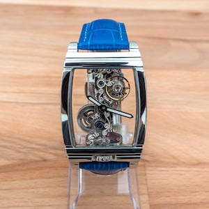 May include: A silver skeleton watch with a blue leather band. The watch face is clear and shows the intricate inner workings of the watch.