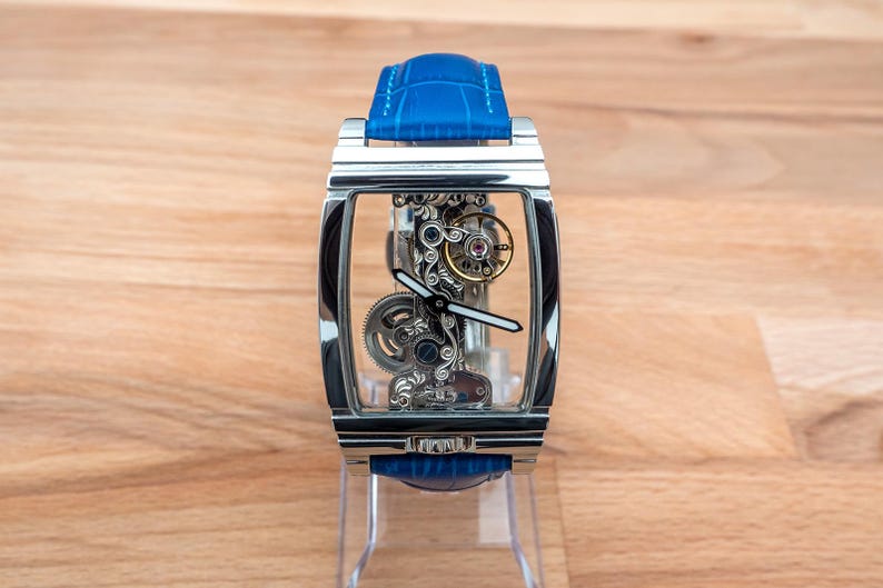 May include: A silver skeleton watch with a blue leather band. The watch face is clear and shows the intricate inner workings of the watch.