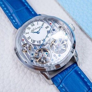 May include: A silver-toned, skeletonized wristwatch with a blue leather band. The watch face features Roman numeral hour markers and a white dial with the word "Ailang" printed on it.