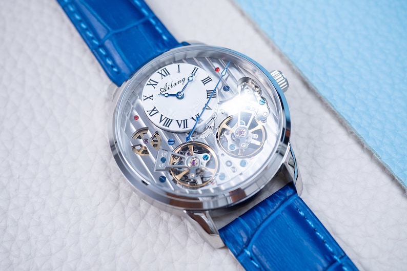 May include: A silver-toned, skeletonized wristwatch with a blue leather band. The watch face features Roman numeral hour markers and a white dial with the word "Ailang" printed on it.