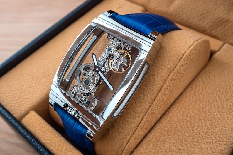 May include: A silver skeleton watch with a blue leather band, displayed in a brown leather watch box.