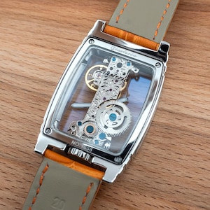 May include: A rectangular, silver-toned wristwatch with a transparent face, showcasing the intricate internal mechanisms. The watch has a light gray leather strap with orange accents. The watch is set against a wooden surface.