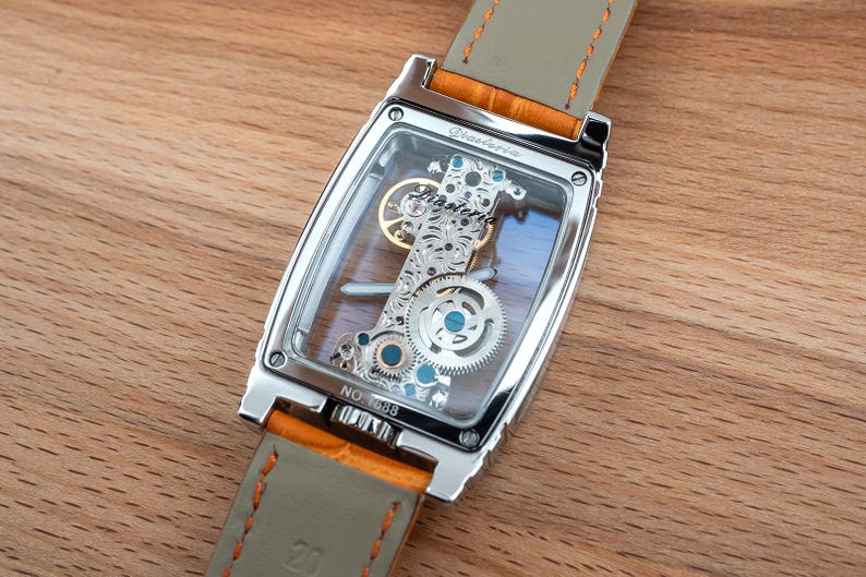 May include: A rectangular, silver-toned wristwatch with a transparent face, showcasing the intricate internal mechanisms. The watch has a light gray leather strap with orange accents. The watch is set against a wooden surface.