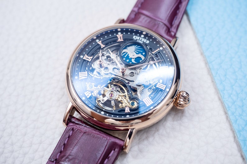 May include: A rose gold skeleton watch with a black face and a burgundy leather strap. The watch face features Roman numeral hour markers and a moon phase indicator. The text "KINYUED 1986" is printed on the watch face.