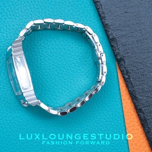 Puede incluir: Un reloj plateado con una esfera rectangular y una correa de metal plateado. El reloj est&aacute; sobre un fondo azul y naranja.