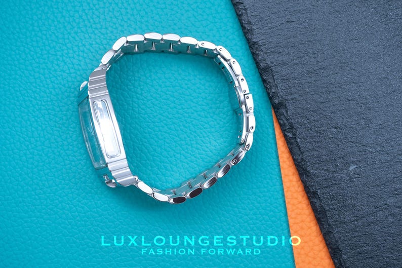 Puede incluir: Un reloj plateado con una esfera rectangular y una correa de metal plateado. El reloj est&aacute; sobre un fondo azul y naranja.