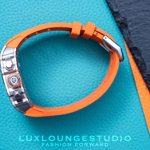 Peut inclure: Gros plan d'une montre-bracelet avec un bracelet orange et un cadran argent&eacute;. La montre est pos&eacute;e sur un fond bleu textur&eacute;. Le texte "LUXLOUNGESTUDIO FASHION FORWARD" est visible en bas.