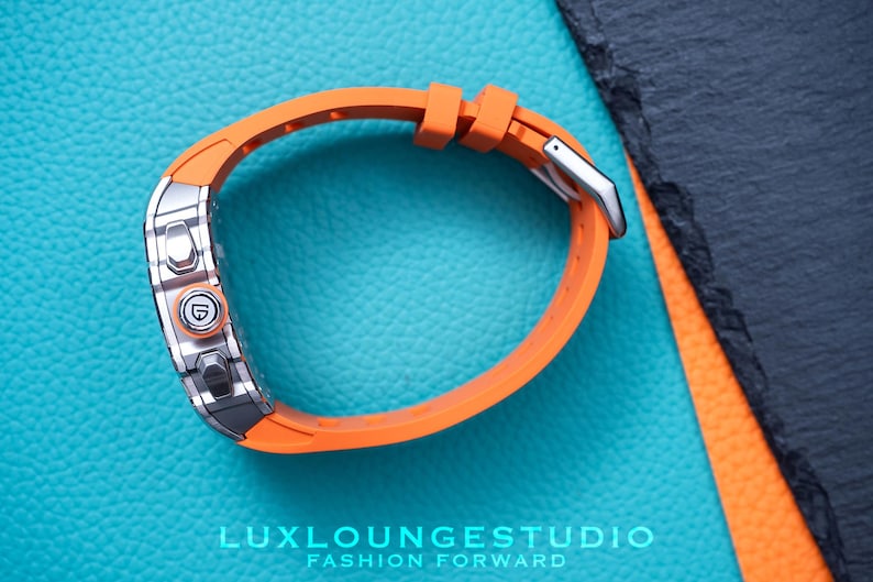 Peut inclure: Gros plan d'une montre-bracelet avec un bracelet orange et un cadran argent&eacute;. La montre est pos&eacute;e sur un fond bleu textur&eacute;. Le texte "LUXLOUNGESTUDIO FASHION FORWARD" est visible en bas.