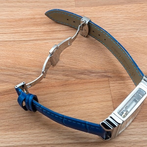 May include: A blue leather watch strap with a silver buckle and a clear case back. The strap is attached to a silver watch case.