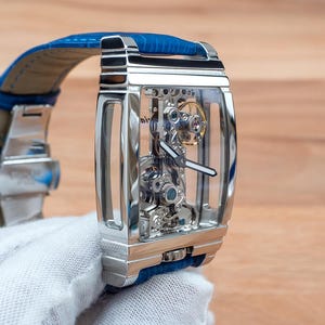 May include: A silver skeleton watch with a blue leather band. The watch face is clear, revealing the intricate inner workings of the watch.
