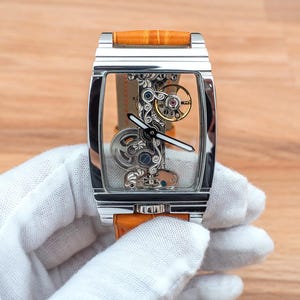 May include: A rectangular, skeletonized wristwatch with a silver case and an orange leather band. The watch face reveals intricate internal mechanisms with gold, silver, and blue components. The watch is held by a gloved hand.