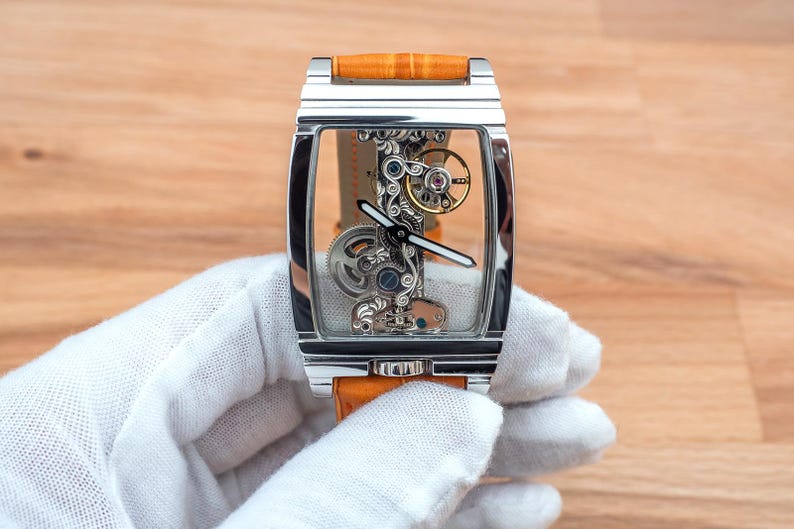 May include: A rectangular, skeletonized wristwatch with a silver case and an orange leather band. The watch face reveals intricate internal mechanisms with gold, silver, and blue components. The watch is held by a gloved hand.