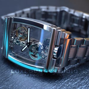 Puede incluir: Un reloj plateado con una esfera transparente y una correa de metal plateado. La esfera del reloj muestra el funcionamiento interno del reloj.