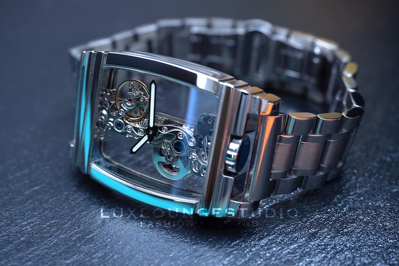 Puede incluir: Un reloj plateado con una esfera transparente y una correa de metal plateado. La esfera del reloj muestra el funcionamiento interno del reloj.