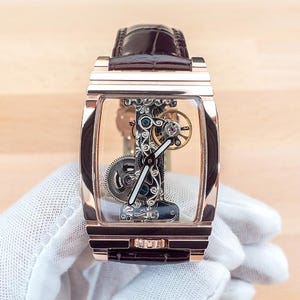 May include: A rose gold rectangular wristwatch with a transparent face, showcasing intricate internal mechanisms. The watch has a brown leather strap and is held by a gloved hand. The watch's gears and components are visible.