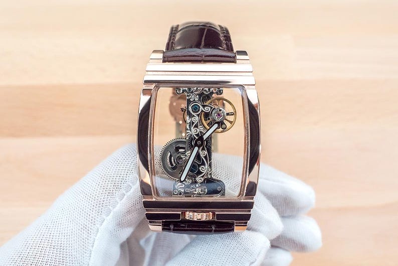 May include: A rose gold rectangular wristwatch with a transparent face, showcasing intricate internal mechanisms. The watch has a brown leather strap and is held by a gloved hand. The watch's gears and components are visible.