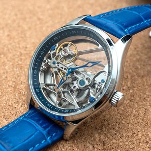 Puede incluir: Un reloj esqueleto plateado con una correa de cuero azul. La esfera del reloj revela el intrincado funcionamiento interno del mecanismo. La correa azul tiene un patrón texturizado similar al de la piel de cocodrilo.