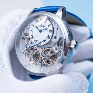 May include: A silver skeleton watch with a white face and blue hands. The watch has Roman numeral hour markers and a blue leather band.