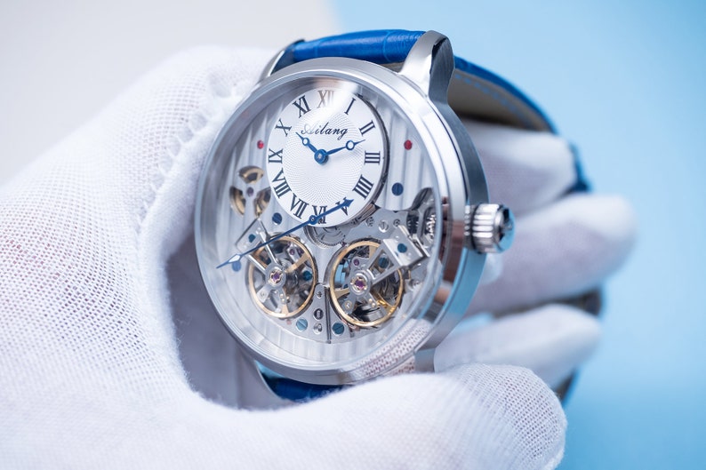 May include: A silver skeleton watch with a white face and blue hands. The watch has Roman numeral hour markers and a blue leather band.