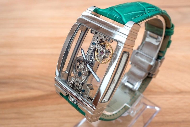 May include: A skeleton watch with a rectangular stainless steel case and a green leather strap. The watch's intricate internal mechanisms are visible through the transparent face. The watch is displayed on a clear stand against a wooden background.
