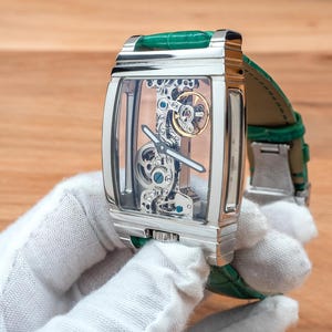 May include: A rectangular, silver wristwatch with a transparent face, revealing the internal mechanisms. It has a green leather strap. The watch is held by a person wearing a white glove. The watch's design is intricate.