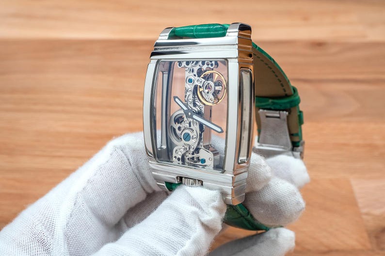May include: A rectangular, silver wristwatch with a transparent face, revealing the internal mechanisms. It has a green leather strap. The watch is held by a person wearing a white glove. The watch's design is intricate.