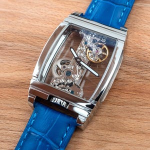 May include: A silver watch with a blue leather band. The watch face is clear and shows the inner workings of the watch.