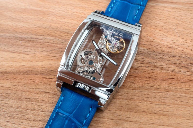 May include: A silver watch with a blue leather band. The watch face is clear and shows the inner workings of the watch.