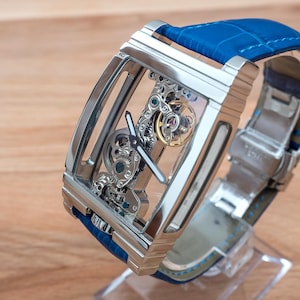 May include: A silver skeleton watch with a blue leather band. The watch face is clear, revealing the intricate inner workings of the watch.