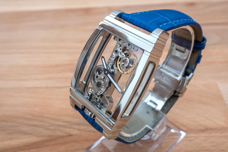 May include: A silver skeleton watch with a blue leather band. The watch face is clear, revealing the intricate inner workings of the watch.