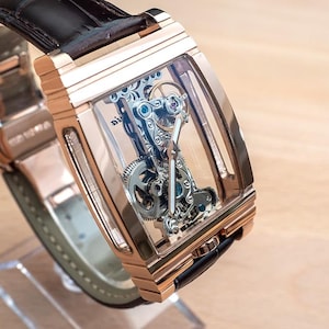 May include: A rose gold rectangular wristwatch with a transparent face, showcasing the intricate internal mechanism. The watch has a black leather strap and is displayed on a clear stand. The watch's design highlights the precision of its internal components.