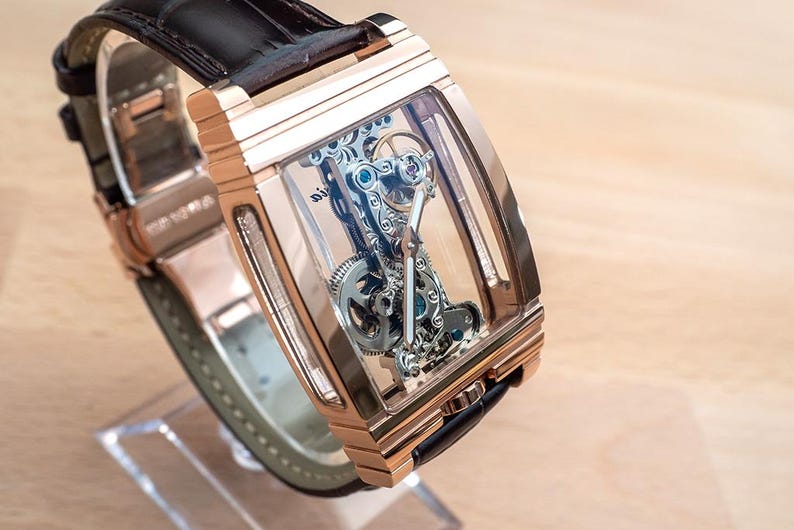 May include: A rose gold rectangular wristwatch with a transparent face, showcasing the intricate internal mechanism. The watch has a black leather strap and is displayed on a clear stand. The watch's design highlights the precision of its internal components.