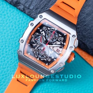 Peut inclure: Une montre de luxe avec un bo&icirc;tier argent&eacute; et un bracelet en silicone orange. Le cadran de la montre pr&eacute;sente un design complexe avec des d&eacute;tails noirs et argent&eacute;s, des accents rouges et des engrenages visibles. La montre est sur une surface bleue textur&eacute;e.