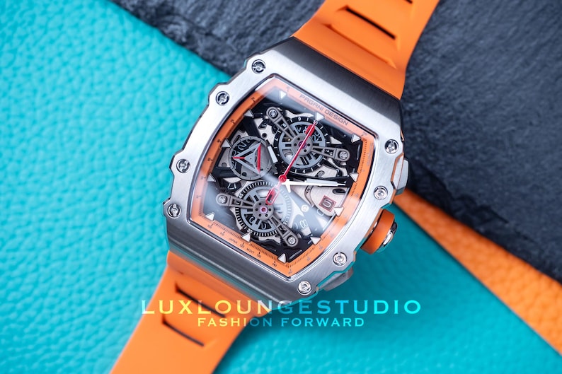 Peut inclure: Une montre de luxe avec un bo&icirc;tier argent&eacute; et un bracelet en silicone orange. Le cadran de la montre pr&eacute;sente un design complexe avec des d&eacute;tails noirs et argent&eacute;s, des accents rouges et des engrenages visibles. La montre est sur une surface bleue textur&eacute;e.