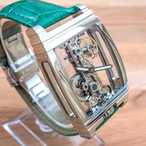 May include: A rectangular, silver wristwatch with a transparent face, revealing the internal mechanisms. It features a green leather strap. The watch is displayed on a clear stand, against a wooden background.
