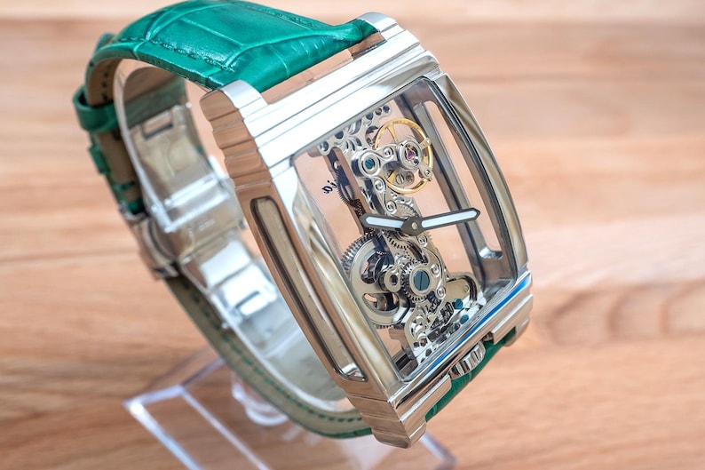 May include: A rectangular, silver wristwatch with a transparent face, revealing the internal mechanisms. It features a green leather strap. The watch is displayed on a clear stand, against a wooden background.