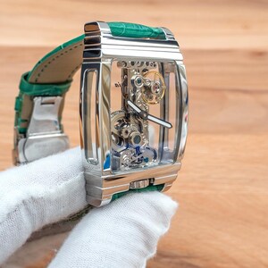 May include: A silver rectangular wristwatch with a transparent face, displaying the internal mechanisms. It features a green leather strap. The watch is held by a gloved hand.